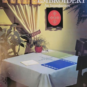 May include: A book titled "Hardanger Embroidery: An Introduction" with a leaflet number of 108 and a price of $2.00. The book cover features a red and black embroidery design on a white background. The book is displayed on a table with a white tablecloth and a green and blue placemat.