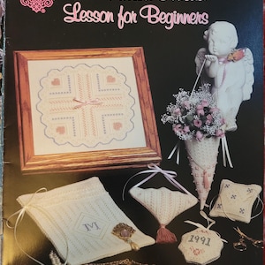 May include: A black background features a framed cross-stitch, a white angel statue, and various needlework projects. The text "A Pulled Thread Lesson for Beginners" is displayed in pink script. Other items include a bag with the letter "M" and a small ornament with "1991".