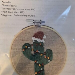 May include: Santa Cactus Ornament embroidery kit. The kit includes a 3-inch embroidery hoop, DMC thread, needle, linen and cotton fabric, felt, and a beginner's guide. The design features a green cactus wearing a Santa hat and string lights.
