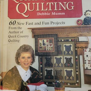 May include: A book cover with the title "More Quick Country Quilting" by Debbie Mumm. The cover features a woman sitting at a table with quilting supplies and a quilt with a bear design.
