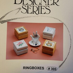 May include: Four small wooden ring boxes with different embroidered designs. The boxes are arranged in a square shape with a white ceramic ring holder in the center. The ring holder is shaped like a hand with four fingers and a thumb. There are three rings on the ring holder. The boxes are labeled "RINGBOXES # 305".