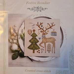 May include: A counted cross stitch pattern titled Samplers and Primitives Festive Reindeer. The design features a brown reindeer with decorated antlers, a green Christmas tree, and small ornaments on a light fabric background with text.