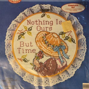 May include: A stamped cross stitch kit featuring a design with the text "Nothing Is Ours But Time." The image shows a woman in a hat holding flowers, stitched in various colors. The kit includes a hoop and is surrounded by a white lace trim.