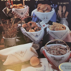 May include: A leaflet titled "BREAD CLOTHS" with nine designs. The image showcases various bread cloths with embroidered designs, displayed in baskets with baked goods. The cloths feature floral and home-themed motifs, with text like "Give us this day our daily bread."