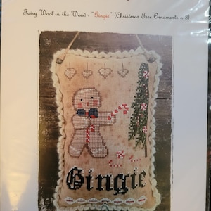 May include: A cross-stitch pattern for a Christmas tree ornament featuring a gingerbread person holding a candy cane and standing next to a small Christmas tree. The pattern is titled "Fairy Wool in the Wood - "Gingie" (Christmas Tree Ornaments n 3)".
