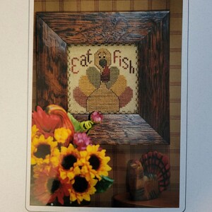 May include: Cross stitch pattern featuring a turkey with the text "Eat Fish". The pattern is framed in a brown wooden frame and surrounded by sunflowers and a wooden turkey figurine. The pattern is from Hillside Samplings.
