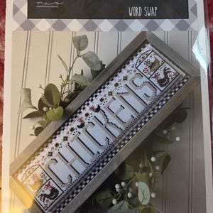 May include: A framed cross-stitch project with the word "CHICKENS" in large letters. The design includes chicken illustrations and decorative patterns. The frame is wooden, and the background is a mix of white and black patterns. The text "WORD SWAP" is also visible.