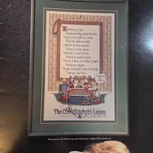 May include: Framed cross-stitch art titled "The CrossStitcher's Legacy." The artwork features a poem about family and cross-stitching, with a scene of children in a bed. The frame is green, and the background is a dark color.