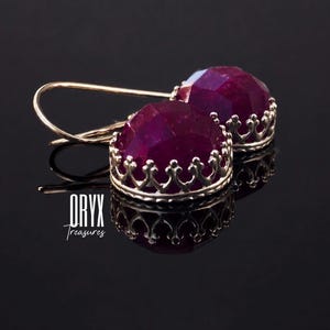 May include: A pair of drop earrings featuring deep magenta, faceted, round gemstones set in silver-toned metal crowns with delicate scalloped edges and curved ear wires. The text ORYX Treasures is printed in white on the lower left side.