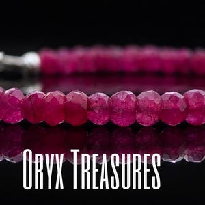 Peut inclure: Un bracelet délicat composé de petites perles de rubis rose foncé à facettes disposées en un brin continu. La pièce présente un mécanisme de fermoir en métal argenté poli à une extrémité. Le texte ORYX TREASURES est visible sous le bijou.