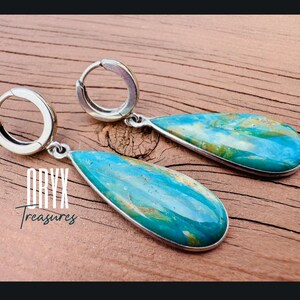 Puede incluir: Pendientes colgantes con forma de l&aacute;grima y marco de metal plateado. Presentan una piedra turquesa y dorada. La imagen incluye el texto "Oryx Treasures".