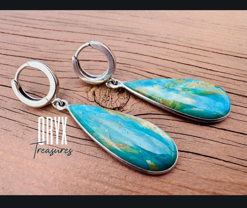Puede incluir: Pendientes colgantes con forma de l&aacute;grima y marco de metal plateado. Presentan una piedra turquesa y dorada. La imagen incluye el texto "Oryx Treasures".