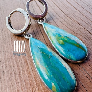 Puede incluir: Pendientes de aro plateados con colgantes en forma de l&aacute;grima de piedras turquesas y azules. Los colgantes presentan un patr&oacute;n veteado con toques amarillos. El texto "ORYX Treasures" es visible.