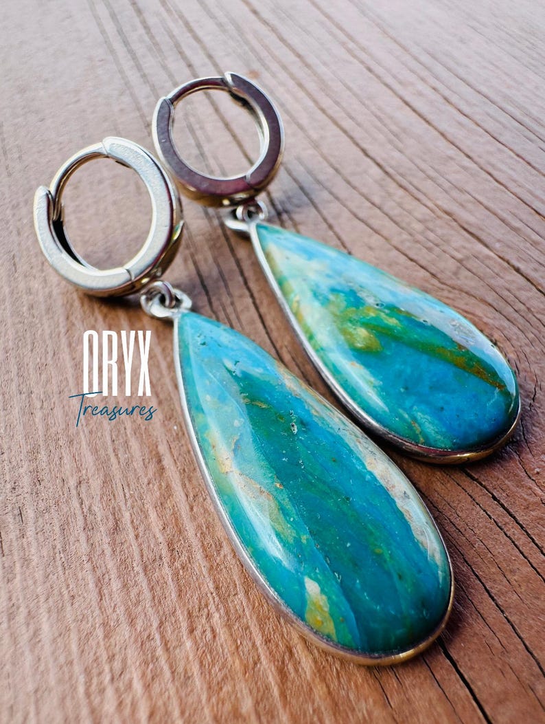 Puede incluir: Pendientes de aro plateados con colgantes en forma de l&aacute;grima de piedras turquesas y azules. Los colgantes presentan un patr&oacute;n veteado con toques amarillos. El texto "ORYX Treasures" es visible.