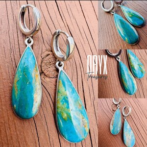 Puede incluir: Pendientes de aro plateados con colgantes de turquesa en forma de l&aacute;grima. La turquesa presenta una mezcla de tonos azules, verdes y marrones. El texto "ORYX Treasures" es visible sobre una superficie de madera.