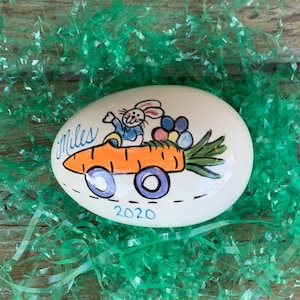 May include: A white ceramic Easter egg with a hand-painted design of a bunny driving a carrot. The bunny is wearing a blue shirt and holding a bouquet of flowers. The egg is decorated with the name "Miles" and the year "2020".