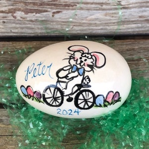 May include: White ceramic Easter egg with a hand-painted design of a bunny riding a bicycle. The bunny is wearing a blue bow tie and the egg is decorated with colorful Easter eggs. The egg is personalized with the name "Peter" and the year "2024".
