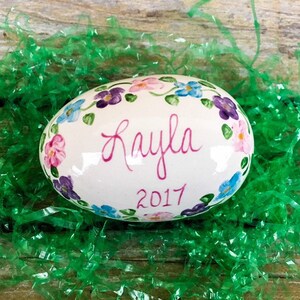 May include: A white ceramic egg with a floral design painted around the top. The egg is decorated with pink, purple, and blue flowers. The name "Kayla" is written in pink cursive on the front of the egg, and the year "2017" is written below it.