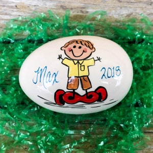 May include: White ceramic egg-shaped ornament with a hand-painted cartoon of a boy riding a hoverboard. The boy is wearing a yellow shirt, brown pants, and red shoes. The ornament is decorated with the name "Max" and the year "2018".