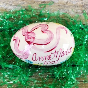 May include: A white ceramic egg with pink ribbon painted on it. The egg is decorated with small yellow and blue flowers. The text "Anne Marie 2002" is written on the egg.