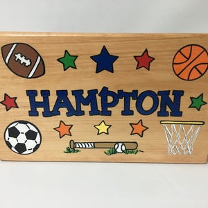May include: Wooden box with hand-painted sports-themed illustrations. Features a football, basketball, soccer ball, baseball bat and ball, and a basketball net. The name "HAMPTON" is prominently displayed in blue letters, surrounded by colorful stars.