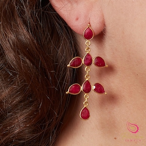 May include: Gold drop earrings with teardrop-shaped, deep red gemstones. The earrings have a tiered design with multiple gemstones suspended from delicate chains. The gemstones are a vibrant, rich red colour, and the earrings are displayed against a skin tone background.
