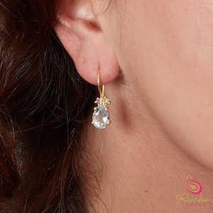 May include: Gold drop earring with a light blue, pear-cut gemstone and small pink and green accent stones. The earring hangs from a gold hook. The Roche Jewels logo is visible in the lower right corner.