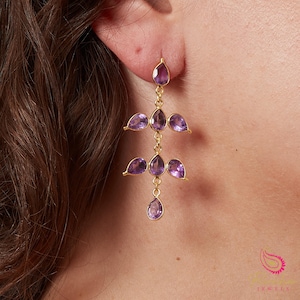 May include: Gold-coloured chandelier earrings featuring teardrop and petal-shaped purple gemstones. The earrings hang from a delicate chain, creating a layered effect. The gemstones are a deep purple colour.