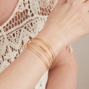May include: A collection of gold-coloured bangles on a wrist. The bangles are slender and delicate, with some featuring small, decorative beads. The person is wearing a white, lace-like top.
