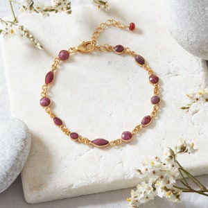 May include: A gold bracelet with a delicate chain and oval-shaped pink gemstones. The bracelet is on a white surface with small white flowers.