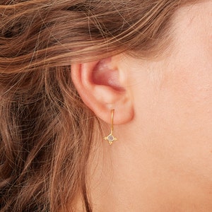 May include: Gold hoop earrings with a small, round, clear gemstone hanging from the bottom. The gemstone is surrounded by three small gold beads.