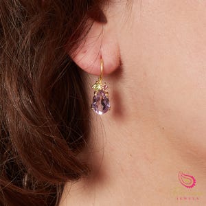 May include: A gold earring with a pear-shaped, pale purple gemstone. The earring has small green and pink stones at the top. The earring is suspended from a person's ear.