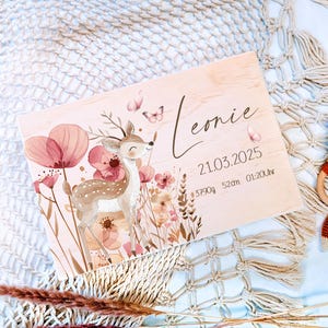 Könnte beinhalten: Holztafel mit Hirsch- und Blumenillustration, dem Namen "Leonie" und Geburtsdaten. Neben der Tafel befindet sich eine gehäkelte Fuchsel-Rassel. Die Tafel zeigt eine Hirschillustration mit rosa Blumen und Schmetterlingen.