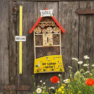May include: A wooden insect hotel with a red roof, measuring 60 cm in height, is mounted on a weathered wooden door. The hotel features various compartments for insects, including bamboo tubes and a wire mesh area. A yellow sign displays the text "Wir ziehen weiter!" and illustrations.