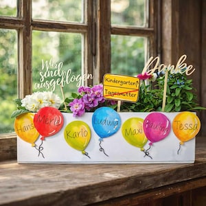 May include: A white wooden planter box with colourful balloon cutouts, each bearing a child's name. The box is filled with flowers and greenery, with wooden signs that read "Danke" and "Wir sind ausgeflogen". A sign says "Kindergarten" and "Tagesmutter".