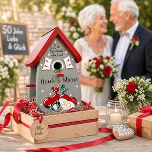 Pode incluir: Uma casinha de pássaros personalizada com telhado vermelho e corpo cinza, decorada com corações, pássaros e os nomes "Heidi & Mario". A casinha está numa caixa de madeira com fita vermelha, ao lado de um biscoito em forma de coração e uma vela acesa, para bodas de ouro.
