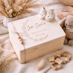 May include: A light wooden keepsake box, engraved with the name "Lea-Sophie" and birth details, including weight and length. The box is decorated with a floral wreath and twine. Baby booties and a wooden toy are also present.