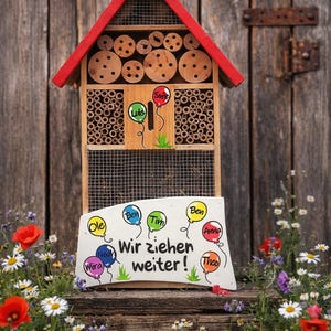 May include: A wooden insect hotel with a red roof and a sign that reads "Wir ziehen weiter!" with names on balloons. The hotel has multiple compartments filled with bamboo and wooden discs. The year "2022" is displayed on top.