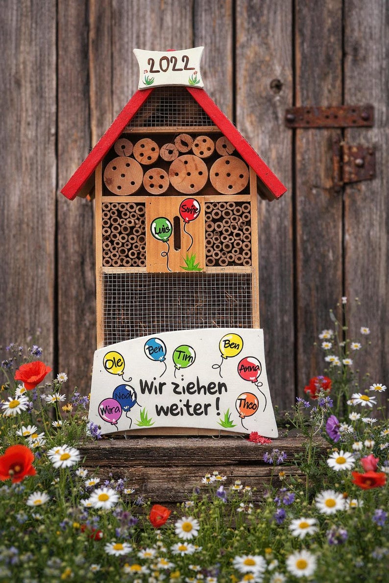 May include: A wooden insect hotel with a red roof and a sign that reads "Wir ziehen weiter!" with names on balloons. The hotel has multiple compartments filled with bamboo and wooden discs. The year "2022" is displayed on top.