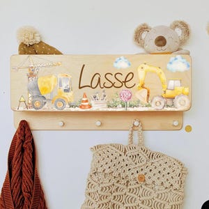 Peut inclure: Organisateur mural en bois sur le thème de la construction, avec une grue, un camion benne et une excavatrice. Le nom "Lasse" est écrit sur l'organisateur. Comprend une étagère et des crochets. Un jouet en peluche koala est posé dessus.