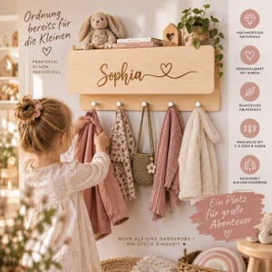 May include: A light-colored wooden wall-mounted coat rack featuring a shelf on top and five silver hooks. The front panel is engraved with the name Sophia and a heart symbol. Various children's clothing items and a small woven bag hang from the hooks.