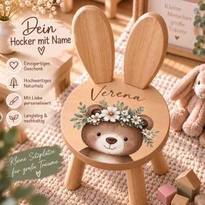 Pode incluir: Um banco de madeira clara com dois encostos altos e arredondados que lembram orelhas de coelho. O assento apresenta uma ilustração de um urso castanho com uma coroa de flores brancas e o nome Verena escrito em letra cursiva.