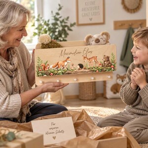 Peut inclure: Une étagère murale en bois clair avec cinq petits crochets, ornée d'une illustration imprimée représentant un renard, un cerf, un écureuil, un hérisson et un ours en forêt, avec le nom Maximilian Elias inscrit en lettres cursives noires.