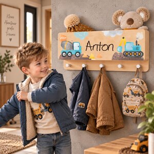 Peut inclure: Un porte-manteau mural en bois clair orné d'un motif de chantier avec une bétonnière bleue, une pelleteuse bleue et le prénom Anton. Le support comporte cinq patères blanches pour suspendre des objets et une étagère supérieure plate.