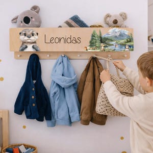 Peut inclure: Un porte-manteau mural en bois avec un nom, une illustration de loutre et un paysage. Le porte-manteau contient une veste bleu marine, un sweat à capuche bleu clair et un manteau marron. Un enfant accroche un sac beige.