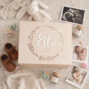 Könnte beinhalten: Eine helle Holzkiste mit dem Namen "Ella" in weißer Schrift, umgeben von einem floralen Kranz. Die Kiste ist von Babyartikeln umgeben, darunter braune Lederschuhe, Schnuller und Fotos. Die Kiste hat auch das Geburtsdatum und die Uhrzeit.