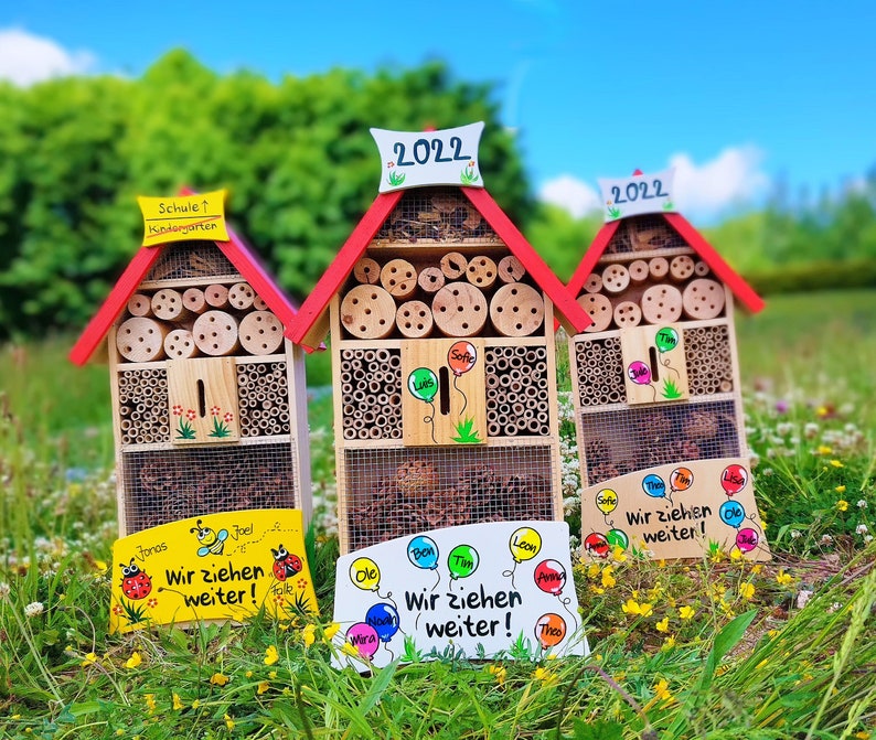 May include: Three wooden insect houses with red roofs and various openings filled with bamboo and wood pieces. Each house has a sign that says "2022" and a sign with the text "Wir ziehen weiter!" and names. The houses are on a grassy lawn.