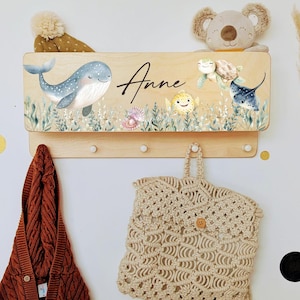 Peut inclure: Porte-manteau mural en bois sur le thème marin. Il présente une baleine, des tortues de mer et d'autres créatures marines. Le nom "Anne" est écrit en cursive. Comprend quatre chevilles en bois pour suspendre des articles. Un pull à capuche marron et un sac en macramé beige sont suspendus en dessous.