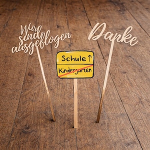 May include: Wooden signs with the phrases "Wir sind ausgeflogen" and "Danke" in cursive script. A yellow sign with "Schule" and "Kindergarten" crossed out is also present. The signs are on wooden sticks, set against a wooden background.