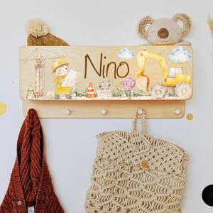 Peut inclure: Porte-manteau mural en bois sur le thème de la construction. Comprend le nom "Nino", un ouvrier de chantier de dessin animé, une excavatrice jaune et un panneau stop. Avec des crochets pour suspendre des articles.